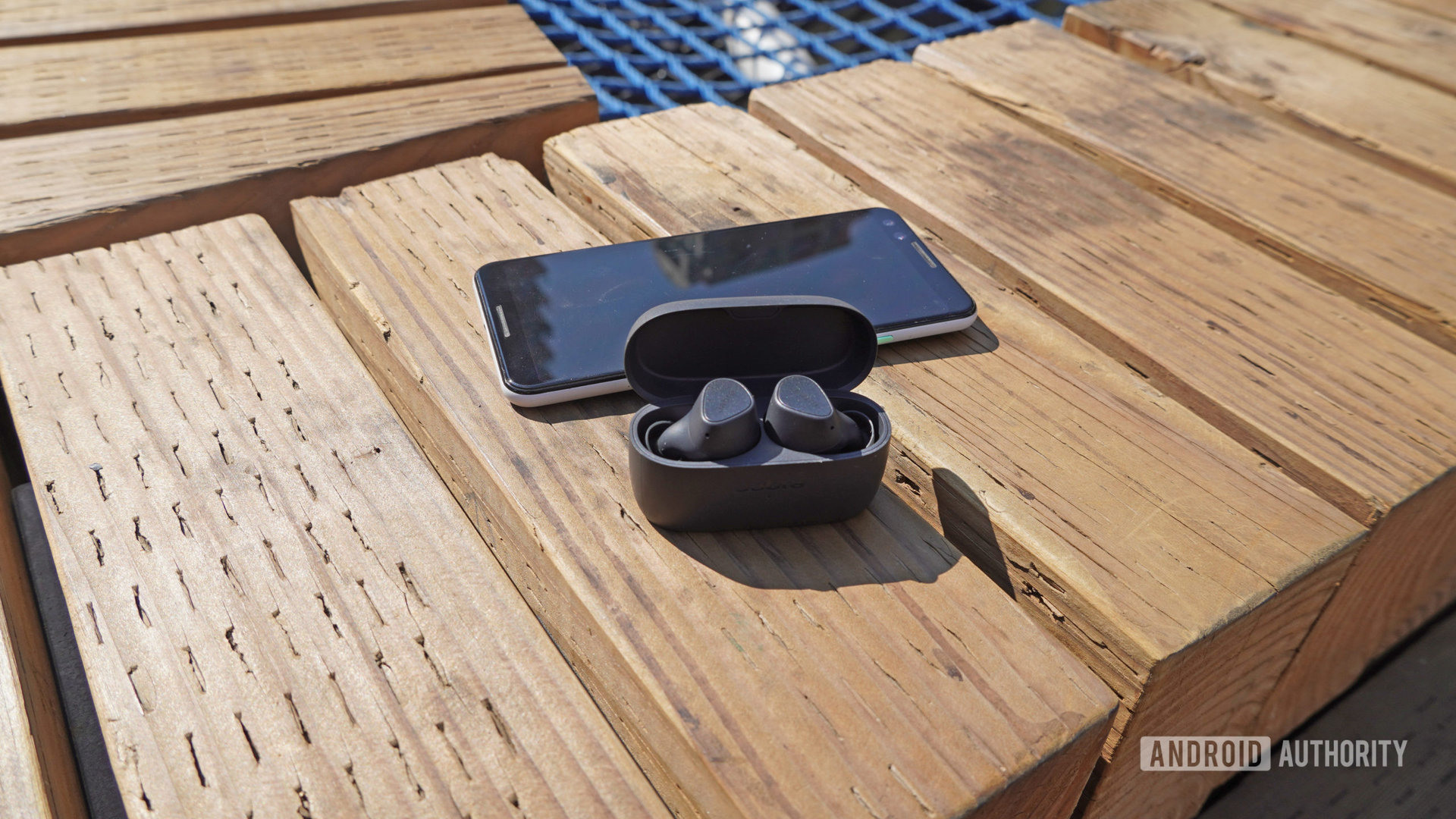 The Jabra Elite 3 earbuds sitting in their case next to a smartphone outdoors on a wooden bench.