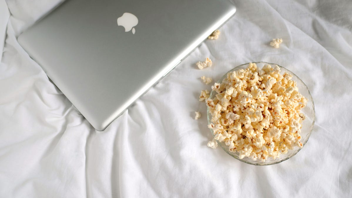 A closed silver MacBook on top of a bed next to a bowl of popcorn.
