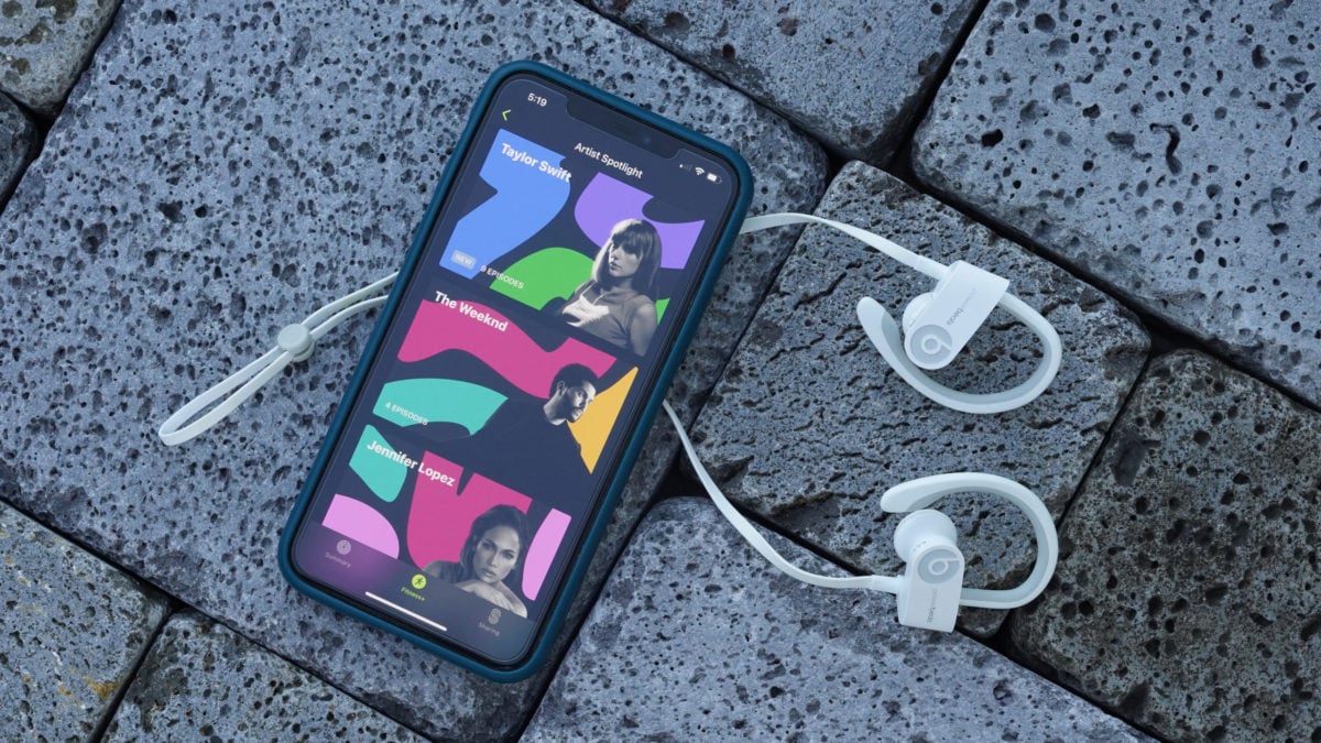 An iPhone displaying Artist Spotlight rests on a brick path along with a pair of wireless headphones.