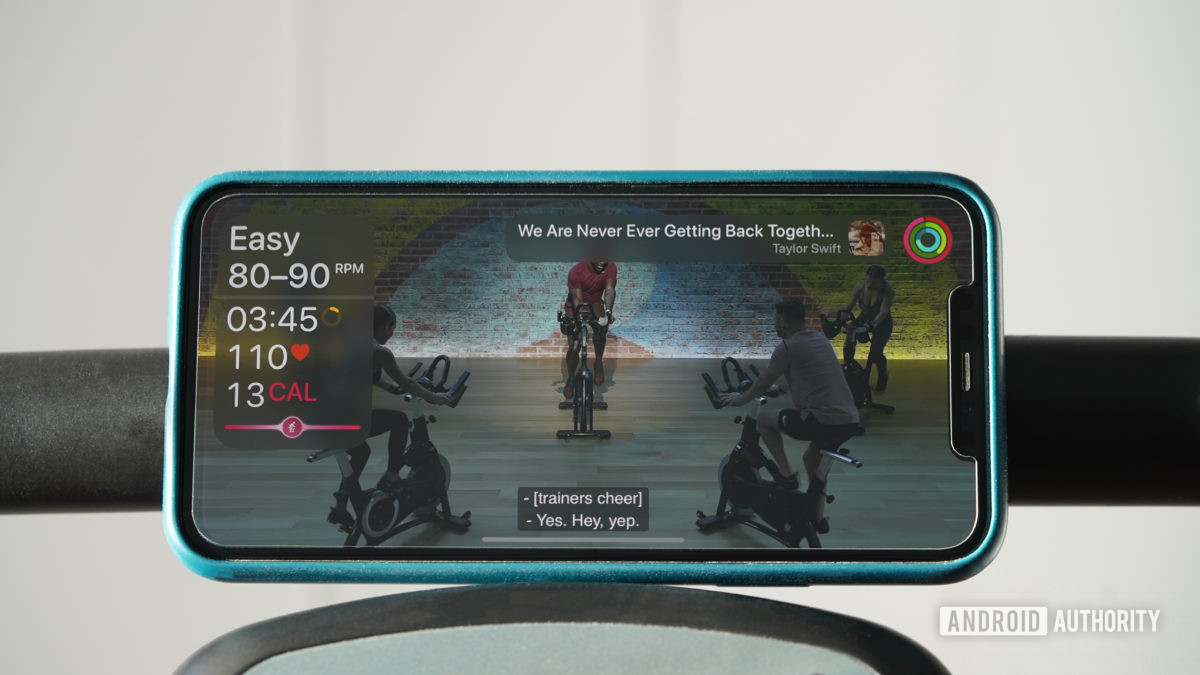 An iPhone balancing on a stationary bike handle displays a cycling workout with the user's live Apple Watch stats displayed.