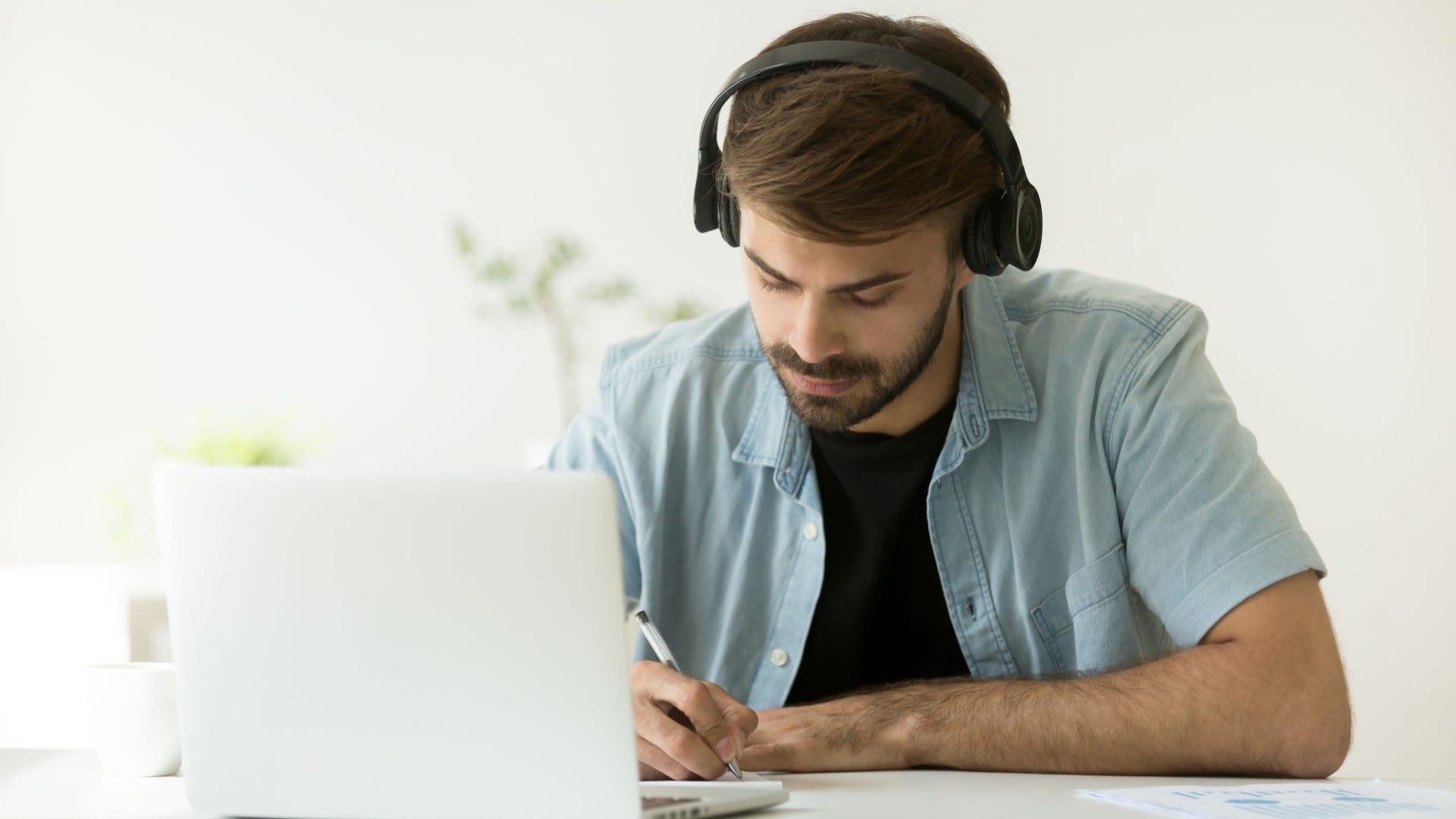 Focused man wearing headphones writing notes studying with laptop.